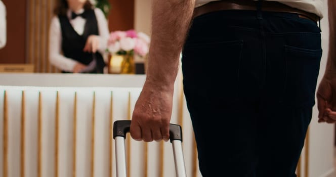 Retired person approaching front desk at hotel, carrying luggage before check in formalities. Tourist talking to employees about all inclusive services, retirement vacation. Close up. Handheld shot.