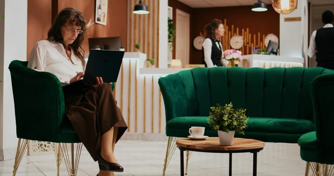 Senior woman using laptop in lobby, navigating internet to look at historical places on online websites. Hotel guest arrives at holiday retreat and waiting for room check in, tourism industry.