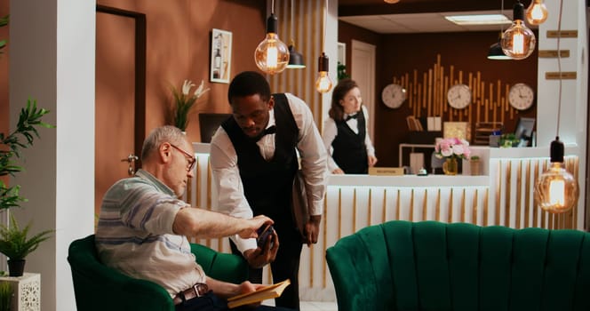 Retired guest paying for coffee using credit card at hotel, ordering beverage from bar and pos terminal payment. Male concierge serving older traveller with refreshment while waiting.