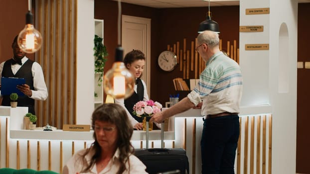 Retired man travelling on vacation, doing check in procedure at front desk in exclusive hotel reception. Concierge assisting elderly tourist with friendly smile while luggage sits nearby.