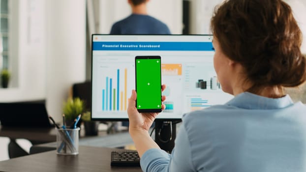 Worker holding device with greenscreen at agency office workstation, startup manager examining isolated mockup template on smartphone. Woman working with blank chromakey layout.