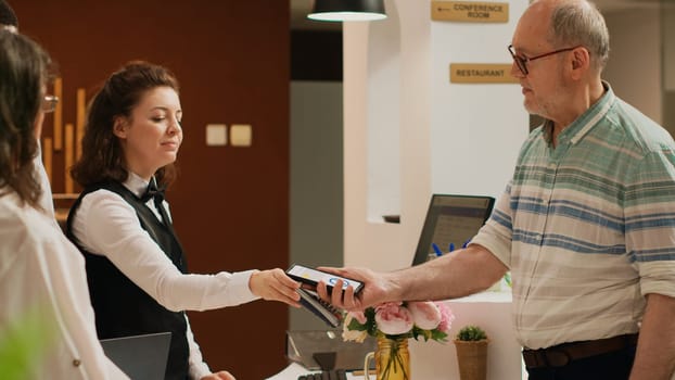 Old tourist using mobile nfc technology to pay for luxury all inclusive hotel room on international vacation. Senior person putting smartphone next to pos terminal, electronic hoiday payment.