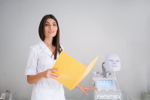 Portrait of smiling young female cosmetologist holding file folder in beauty salon. Woman dermatologist standing near LED mask machine in clinic. Lady professional in white uniform looking at camera.