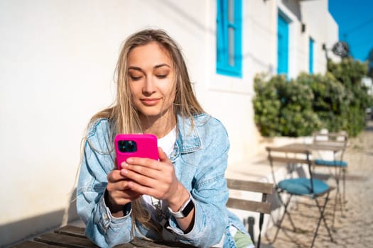 Beautiful blond woman using smartphone at outdoor cafe table. Happy female tourist in denim shirt sitting near white building. Lady enjoys her vacation in typical Portugal city.