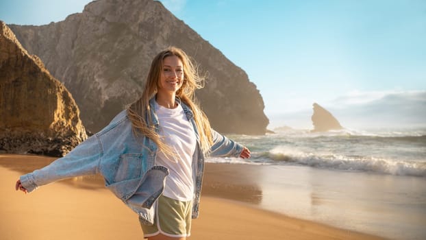 Portrait of smiling blond woman standing arms outstretched on sandy beach. Beautiful female in denim jacket and shorts standing near big rock and sea waves. Lady enjoys summer vacation.