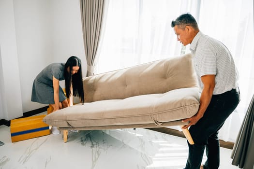 An Asian couple smiles as they move a sofa in their new apartment showcasing relocation teamwork and excitement. Perfect for illustrating furniture replacement and settling into a cozy home.