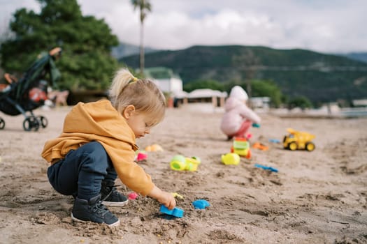 Little girl squats on the beach and plays with plastic sand molds. High quality photo