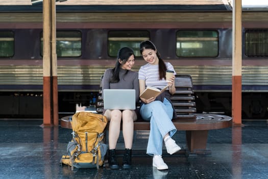 Asian woman friends using laptop and note for planing trip together at railway station have happy moment.