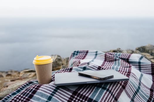 Laptop coffee on blanket with ocean view. Illustrating serene outdoor laptop use. Freelancer enjoying their time outdoors while working or browsing the internet