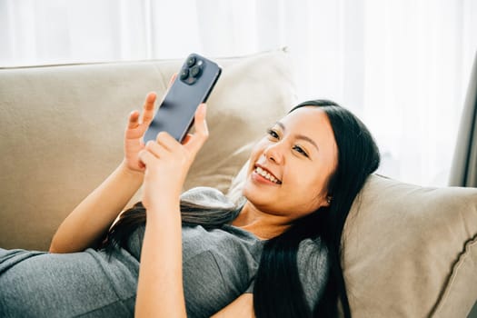 Asian woman on couch texts on smartphone smiling. Enjoying relaxation chatting and online shopping. Modern technology for communication and connection at home.