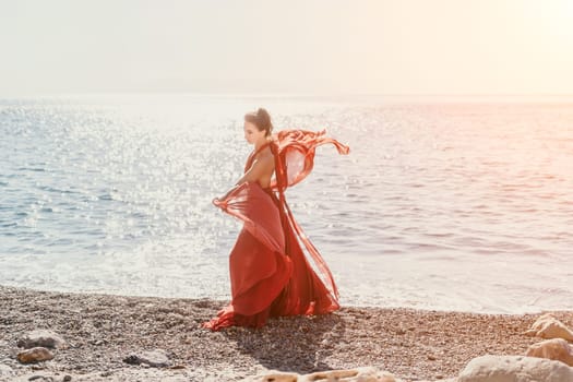 Side view a Young beautiful sensual woman in a red long dress posing on a rock high above the sea during sunrise. Girl on the nature on blue sky background. Fashion photo.