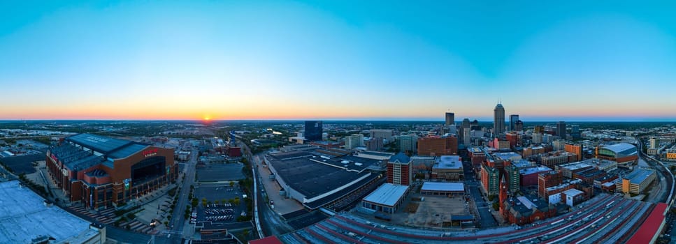 Dusk descends over Indianapolis, showcasing an illuminated Lucas Oil Stadium and vibrant skyline in a serene panorama, captured from an aerial perspective.