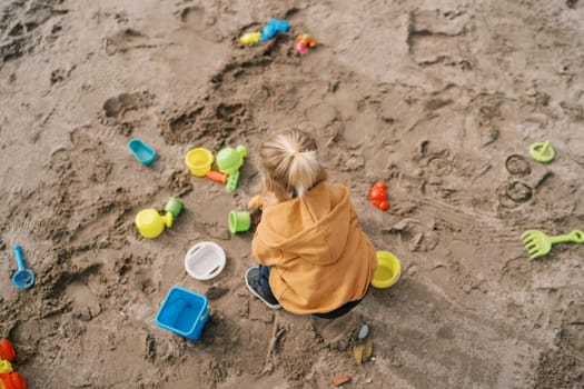 Little girl squats on the sand and plays with colorful toys. Top view. High quality photo