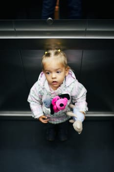 Little girl with soft toys rides in an elevator and looks up. High quality photo
