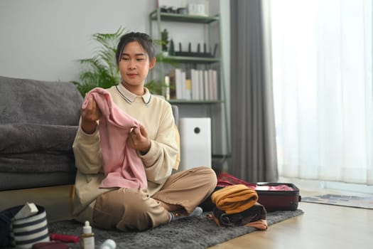 Happy young asian woman packing suitcase, preparing for travel trip at home