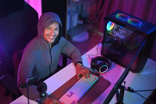 Smiling male gamer in hoodie playing video games on computer at home with neon light.