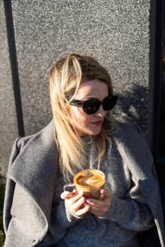 Portrait of tanned lady with cup of latte on foreground. Smiling woman in sunglasses during breakfast in cafe. High quality photo