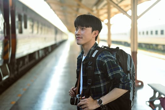 Portrait of happy man tourist with backpack waiting train at railroad station platform.
