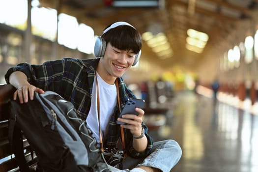 Happy smiling Asian man using mobile phone while waiting train at railroad station platform.