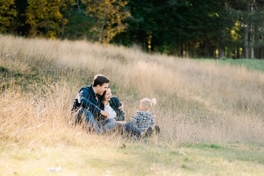 Dad hugs mom sitting in a clearing near a little girl and looking at her. High quality photo