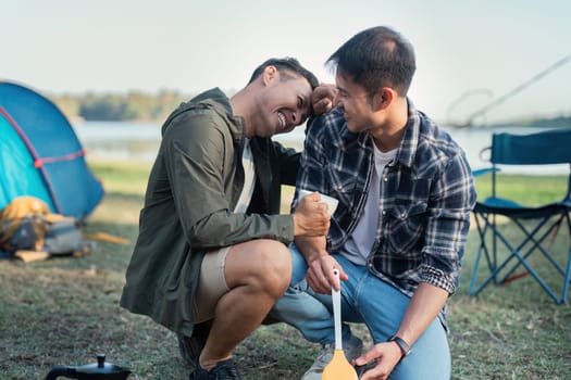Gay LGBTQIA couple sitting on picnic chair drinking tea and making food while camping on vacation holiday.