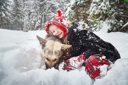 Cute little girl in red cap or hat and black coat with basket of green fir branches in snow forest and big dog shepherd looking as wolf on cold winter day. Fun and fairytale on photo shoot