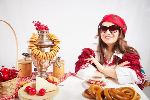 A fashionable modern girl in stylized folk clothes at table with a samovar, bagels and tea for the Orthodox holiday of Maslenitsa and Easter. Funny photo shoot for a young woman