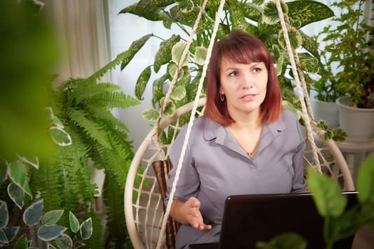 A girl with a laptop and notebook is working on a swing chair in the winter garden. A middle-aged business woman. Psychologist in the office with flowers