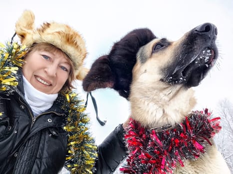 Funny Adult girl with shepherd dog in tinsel, fur caps taking selfie in a winter cold nature. Middle aged woman, big pet and Christmas or New Year