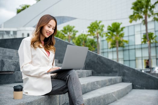 Happy young Asian woman freelance looking laptop with smile while working with coffee cup while sitting on stairs outdoors in the city. Remote working concept in the office.