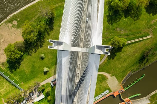Cable-stayed pylon bridge above scenic urban parks with green grass on sunny spring day. Vehicles drive on Redzinski Bridge in Wroclaw upper aerial view