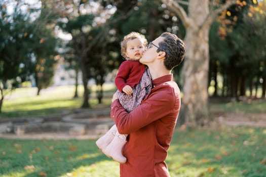 Dad kisses on the cheek a little girl in his arms while standing in a sunny park. High quality photo