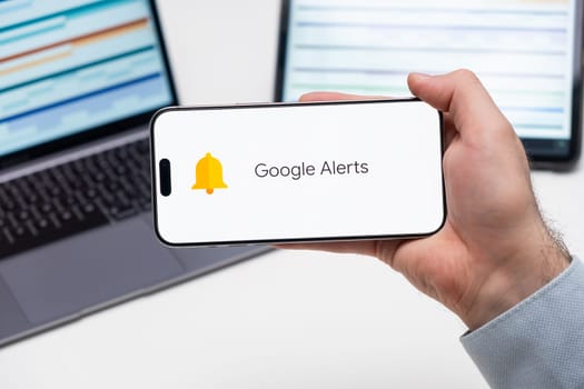 Google Alerts application logo on the screen of smart phone in mans hand, laptop and tablet are on the table in the background, December 2023, Prague, Czech Republic.