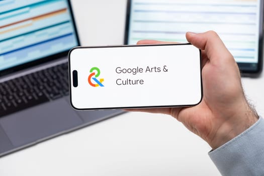 Google Arts and Culture application logo on the screen of smart phone in mans hand, laptop and tablet are on the table in the background, December 2023, Prague, Czech Republic.