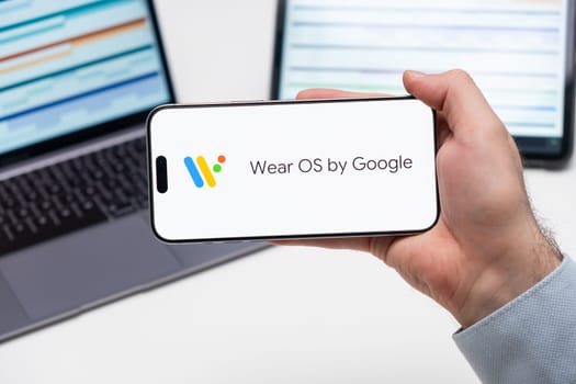 Wear OS by Google application logo on the screen of smart phone in mans hand, laptop and tablet are on the table in the background, December 2023, Prague, Czech Republic.