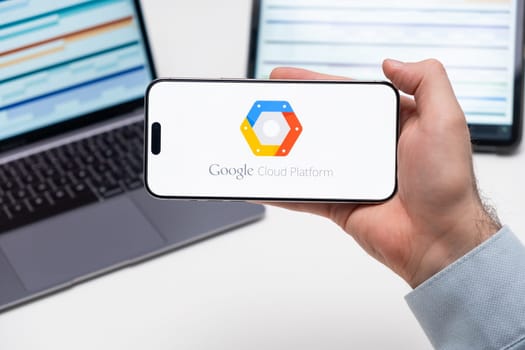 Google Cloud Platform logo of app on the screen of mobile phone held by man in front of the laptop and tablet, December 2023, Prague, Czech Republic