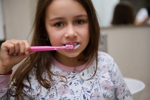 Close-up portrait of a Caucasian school child, adorable little kid girl brushing teeth, looking at the camera. Dental health and oral hygiene concept. People and healthy lifestyle
