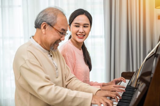 Family care. Happy daughter and elderly father playing music on the piano together in living room, Asian young woman teaching piano for senior man teaching at home, lifestyle life after retirement