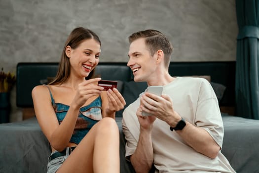 Young couple sit in the home bedroom using online payment app and digital wallet on smartphone to pay with credit card. E commerce shopping and modern purchasing via mobile internet. Unveiling