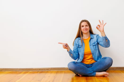 Relaxed woman sitting on a wooden floor, pointing to the side with one hand and making an 'OK' sign with the other, positive gesture.