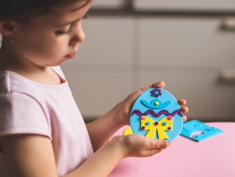A little caucasian girl is holding a blue felt sticker Easter egg in her hand, showing the end result of her needlework, sitting at a children's table with depth of field, close-up side view. Concept of crafts, diy, needlework, artisanal, children art, easter preparation, children creative.