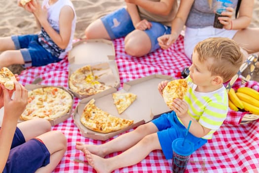 Group of people sitting on a checkered blanket at the beach, sharing multiple pizza boxes and enjoying a casual outdoor meal together.