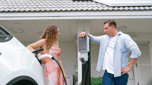 Happy and lovely couple with eco-friendly conscious recharging electric vehicle from EV charging station. EV car technology utilized as alternative transportation for future sustainability. Synchronos