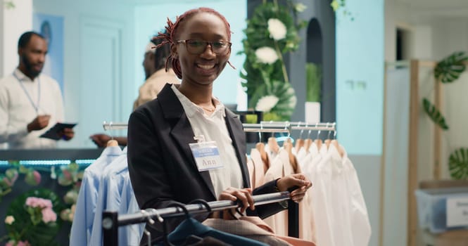 Portrait of woman restocking elegant selection of shirts on hangers in high end fashion boutique. Happy worker arriving with trendy, stylish garments from new collection in clothing shop