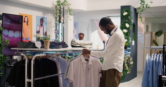 Retail clerk working in luxury fashion boutique, arranging clothes on hangers. African american employee arriving with new stock, showcasing collection of trendy garments on racks