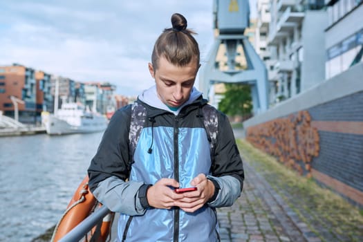 Fashionable young male with backpack using smartphone outdoors in city. Hipster with trendy hairstyle beard with phone in hands mobile applications for tourism travel communication entertainment study