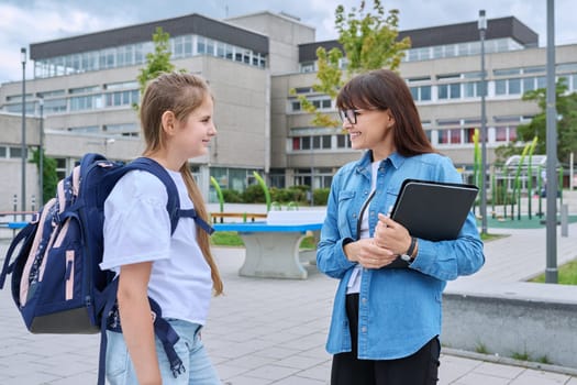 Talking female teacher and schoolgirl child outdoor, school building background. Meeting communication student girl with backpack and mentor counselor. Education, pre-teenage, learning, back to school