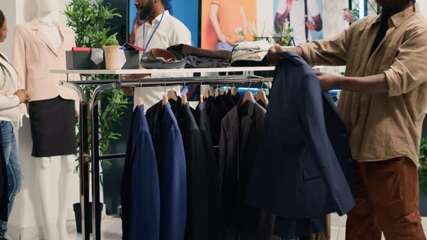 Man looking at elegant blazers in clothing store, trying to determine if materials are qualitative. Client analyzing garments on fashion boutique racks, deciding to buy after finding top quality apparel