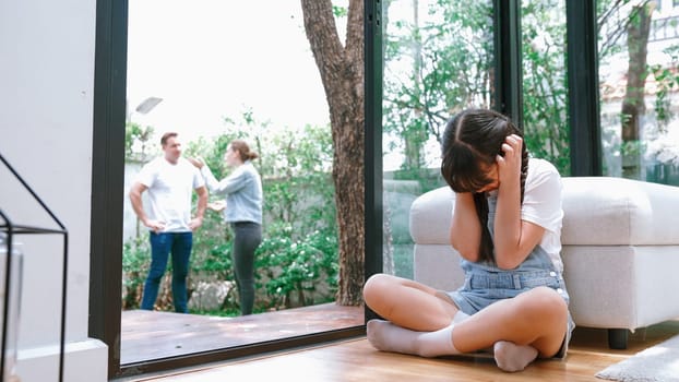 Stressed and unhappy young girl huddle in corner, cover her ears blocking sound of her parent arguing in background. Domestic violence at home and traumatic childhood develop to depression. Synchronos