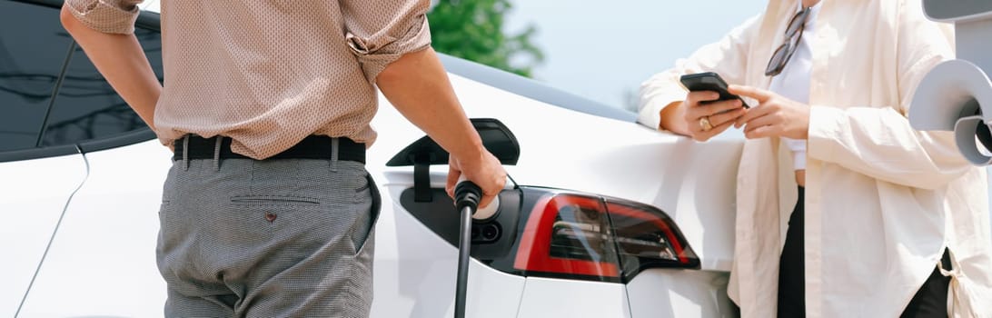 Young couple use smartphone to pay for electricity at public EV car charging station green city park. Modern environmental and sustainable urban lifestyle with EV vehicle. Panorama Expedient